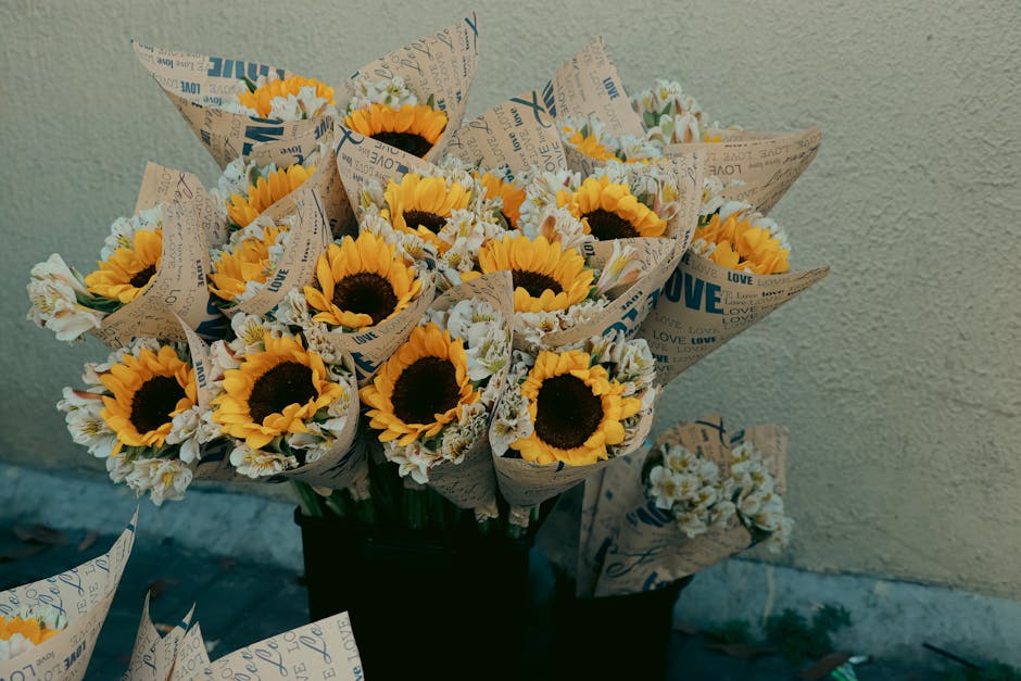 A bouquet arrangement of fresh sunflowers with bright yellow petals and dark centers, complemented by small white daisy-like flowers, all wrapped in patterned paper with words like 'LOVE' and 'LOVE' p